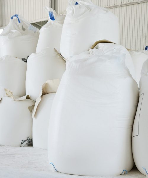 Heaps of huge white sacks containing loose raw materials prepared for production process in spacious warehouse or storage room